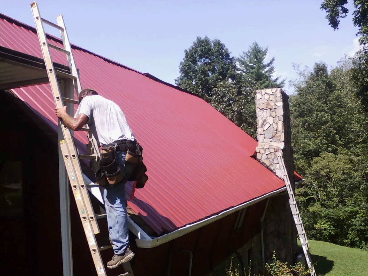 Roofer on a ladder beside a red metal roof and stone chimney — Soffit & Fascia Repair by Roofing Brothers in Peshtigo